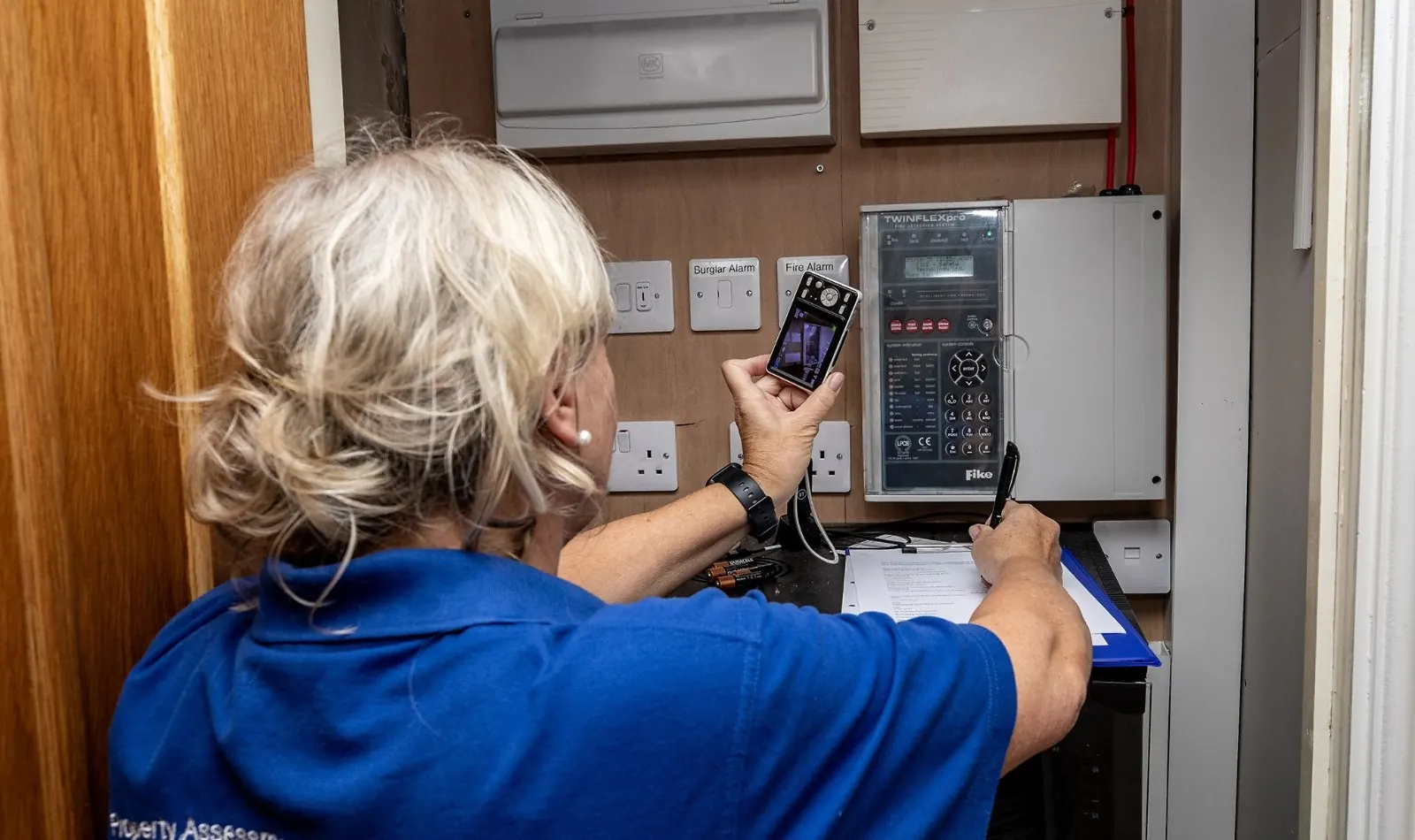 A PALS assessor photographing a fire alarm control panel and making notes on a clipboard