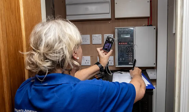 A PALS assessor photographing a fire alarm control panel and making notes on a clipboard