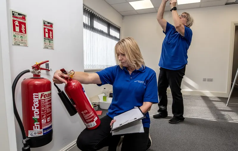 Two PALS assessors working in an office, one checking a foam extinguisher and one inspecting a ceiling-mounted detector